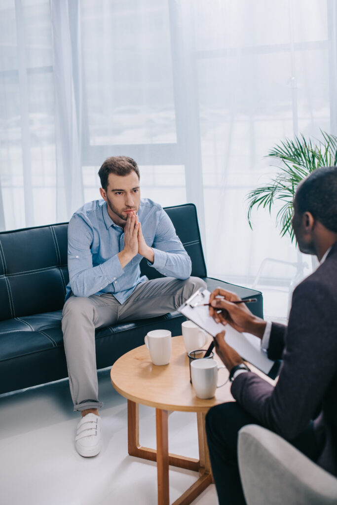 Man with hands pressed together sitting on a black sofa as a counselor takes notes