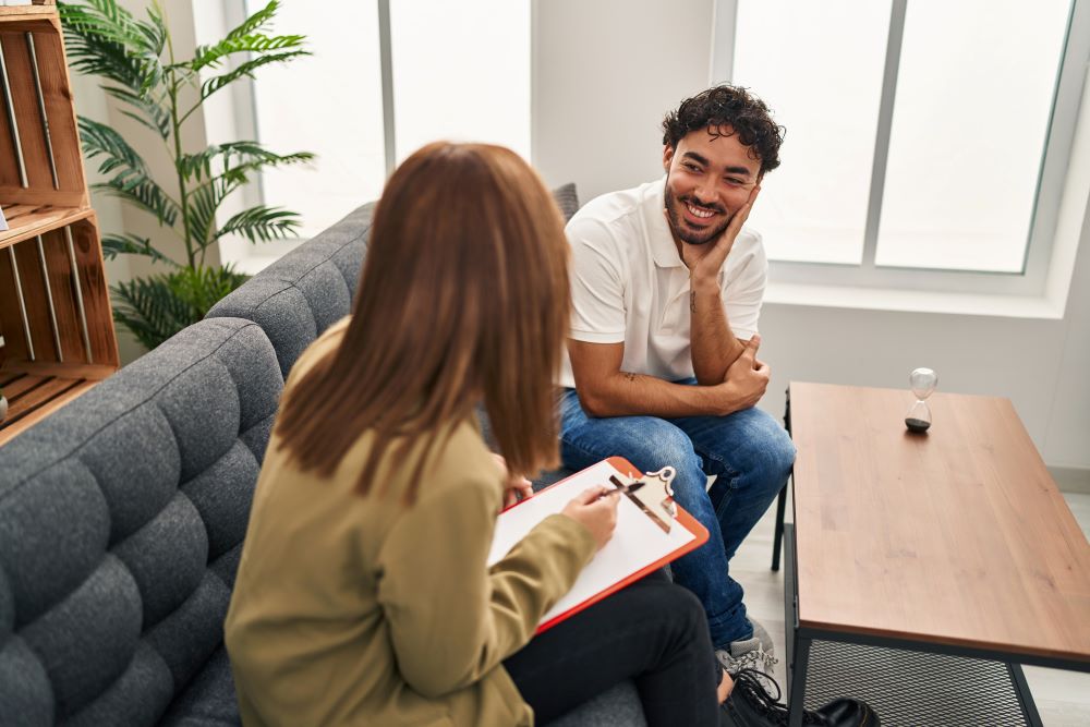 A smiling man sits across from a counselor with a clipboard in a bright therapy room